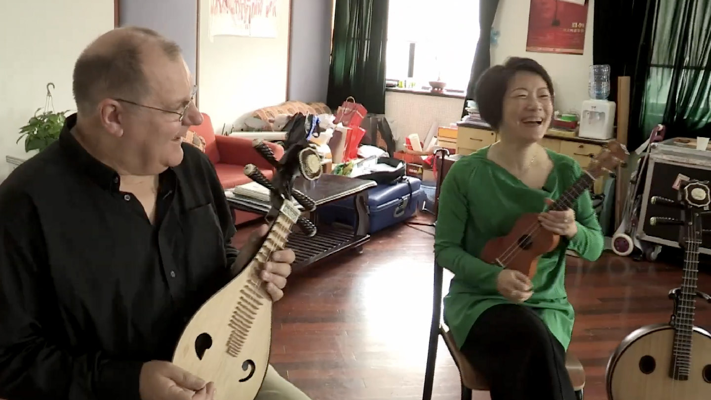 Watch ‘UK Rock God Meets Chinese Axe Goddess’. (George Hinchliffe seated next to Mme. Wu in her studio. George holds a liuqin, Mme. Wu holds a ukulele. They are laughing as they play.)